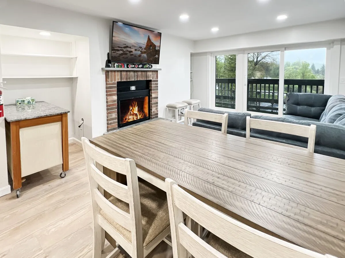 A warm, inviting living/dining area featuring a dining table, a cozy electric fireplace, television, and large sliding glass doors. The space is arranged for family gatherings or group relaxation after a day on the slopes.