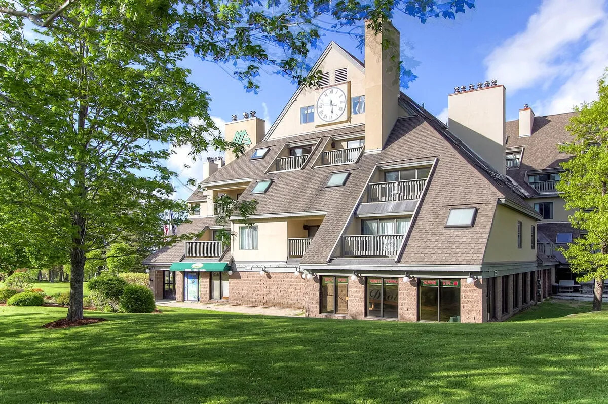 The exterior scene showcases the amenity center's iconic clock tower, expansive green lawn, and surrounding trees. The large windows hint at vibrant indoor amenities, while inviting outdoor spaces encourage gatherings and relaxation in a resort-like setting.