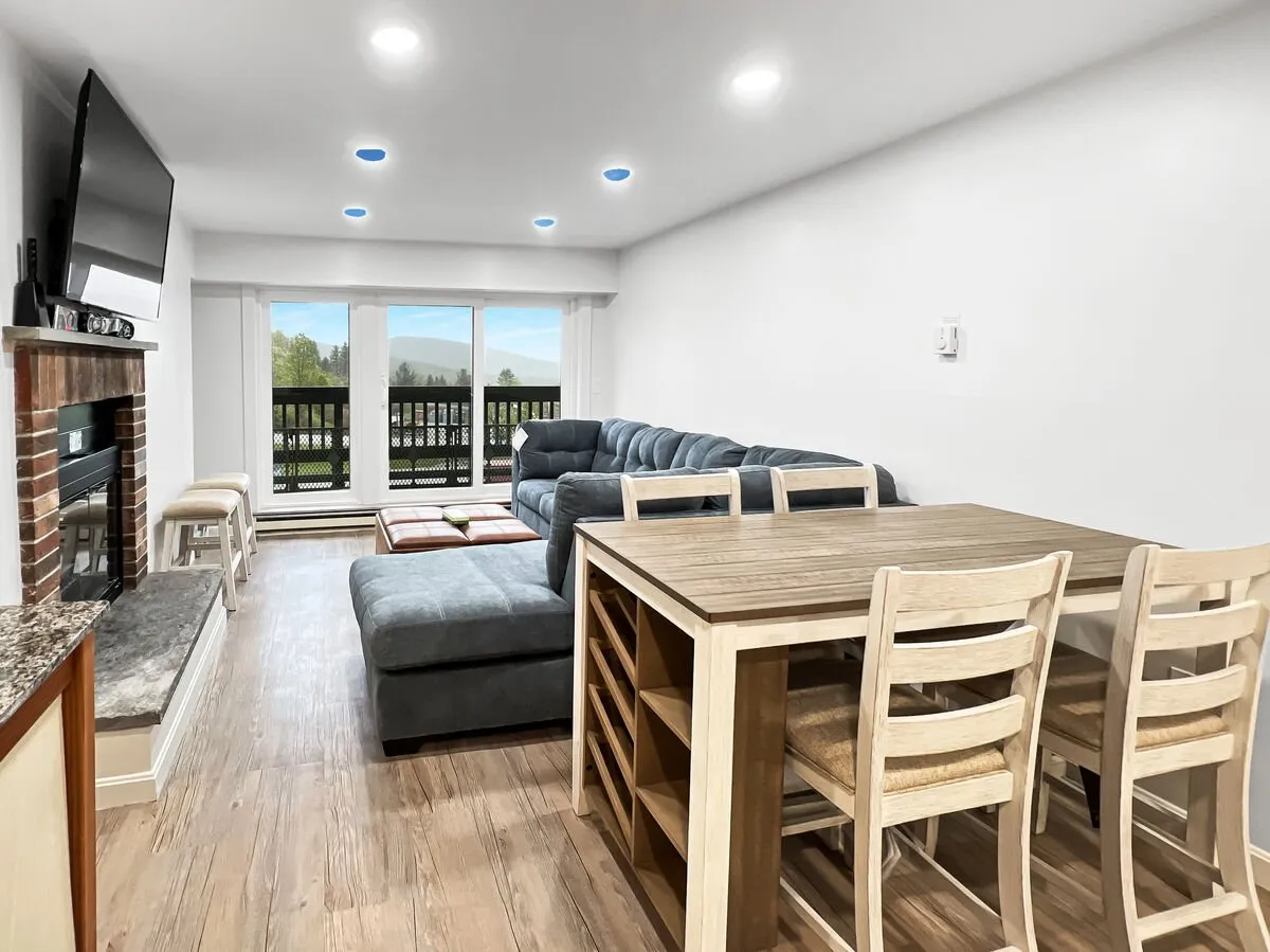 This image beautifully captures the heart of the open-concept living space, showcasing a plush sectional sofa facing a glowing fireplace and a spacious dining table. Large windows bathe the area in natural light, adding warmth and a welcoming ambiance. The scene suggests a space perfect for family gatherings, cozy evenings, and shared meals, aligning perfectly with the section's emphasis on connection and relaxation.