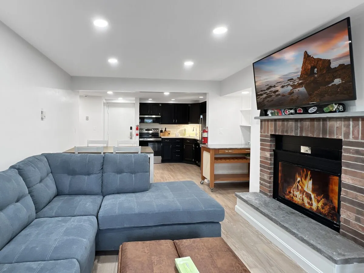 The image features a stylish open-concept living area with a plush sofa set beside a modern fireplace, a wall-mounted television, and a sleek kitchen area—all bathed in warm, inviting light. The setting exudes a sense of comfort and modern elegance, perfect for relaxing or entertaining after a day on the slopes.