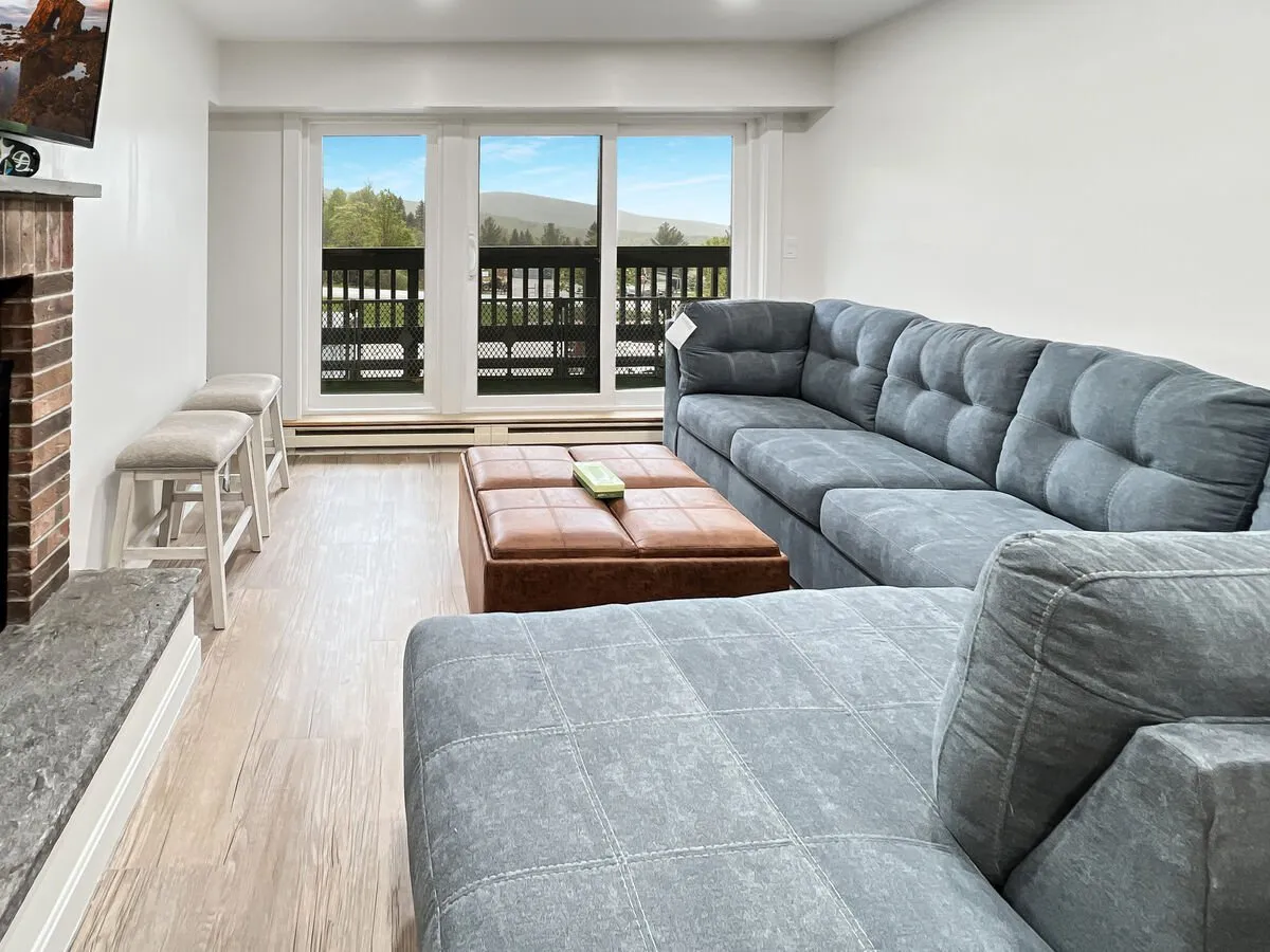 The scene centers on a cozy sectional sofa and coffee table positioned near a glowing fireplace, with expansive windows offering views of the mountains. The layout invites relaxation and socializing, whether for movie nights or watching sunsets. The room’s stylish touches blend comfort with a hint of mountain charm.