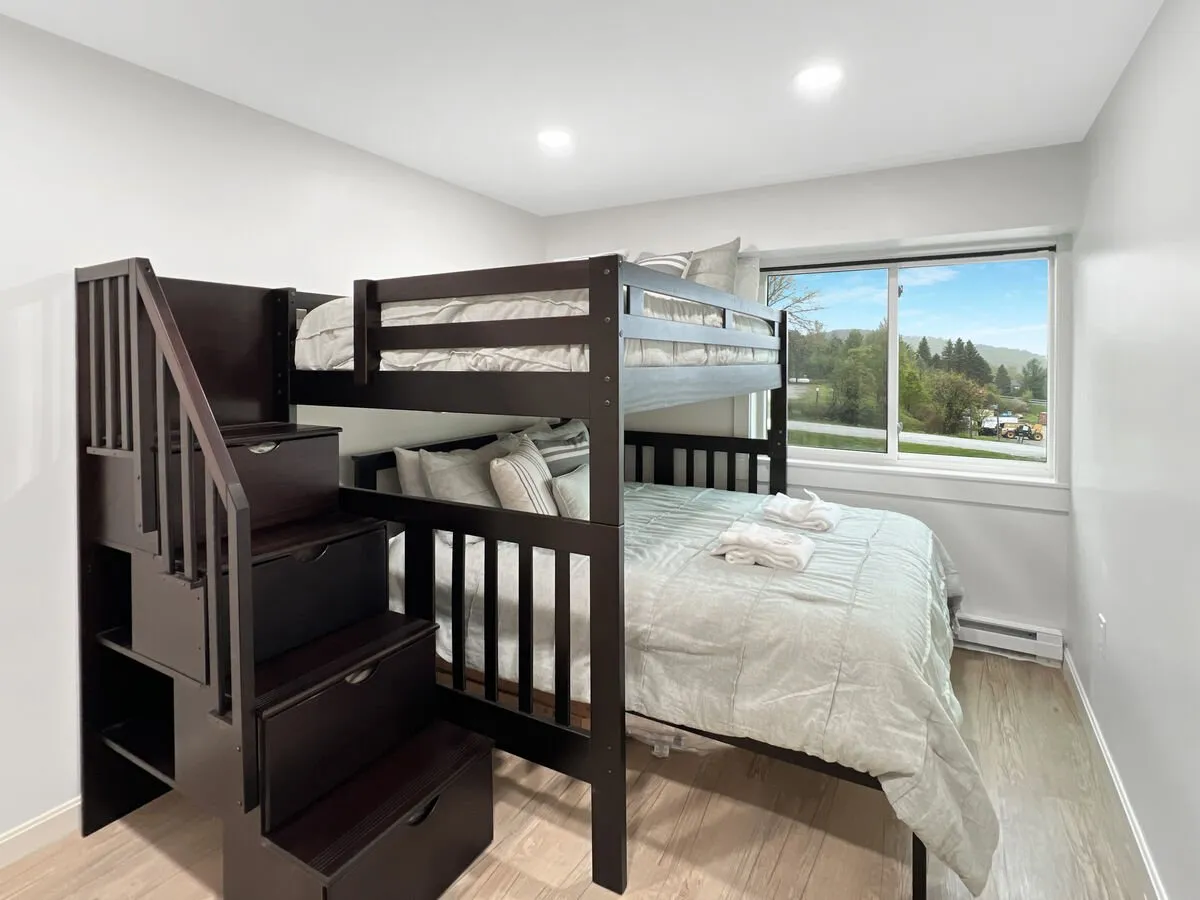This image captures a bright and inviting bedroom featuring a custom bunk bed with a twin above and queen below. The space is illuminated by natural light streaming in from a window, while a compact staircase leads to the upper bunk. The bedding appears fresh and comfortable, and the room feels thoughtfully organized for both children and adults.