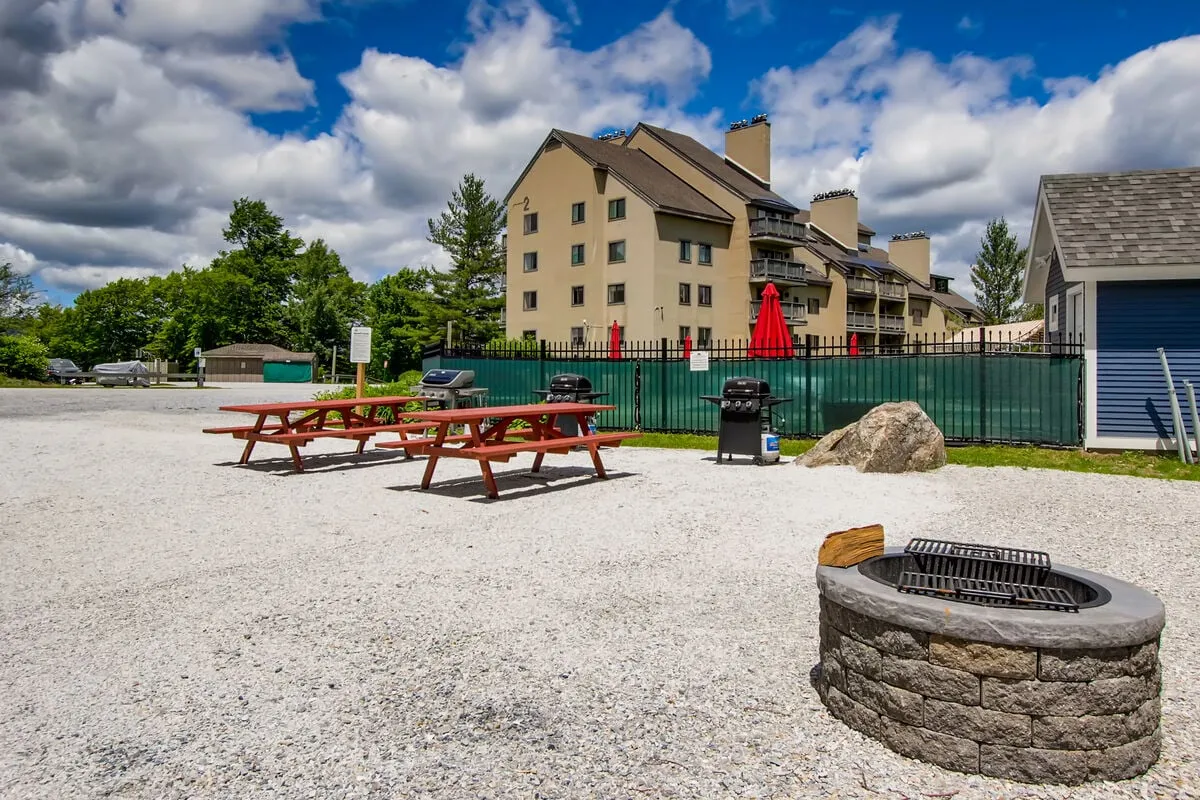 This exterior image features a lively communal area equipped with picnic tables, a fire pit, a grill, and red umbrellas. The building with balconies overlooks the space, and there are groups of people gathered, suggesting an atmosphere perfect for socializing, outdoor dining, and evening relaxation.